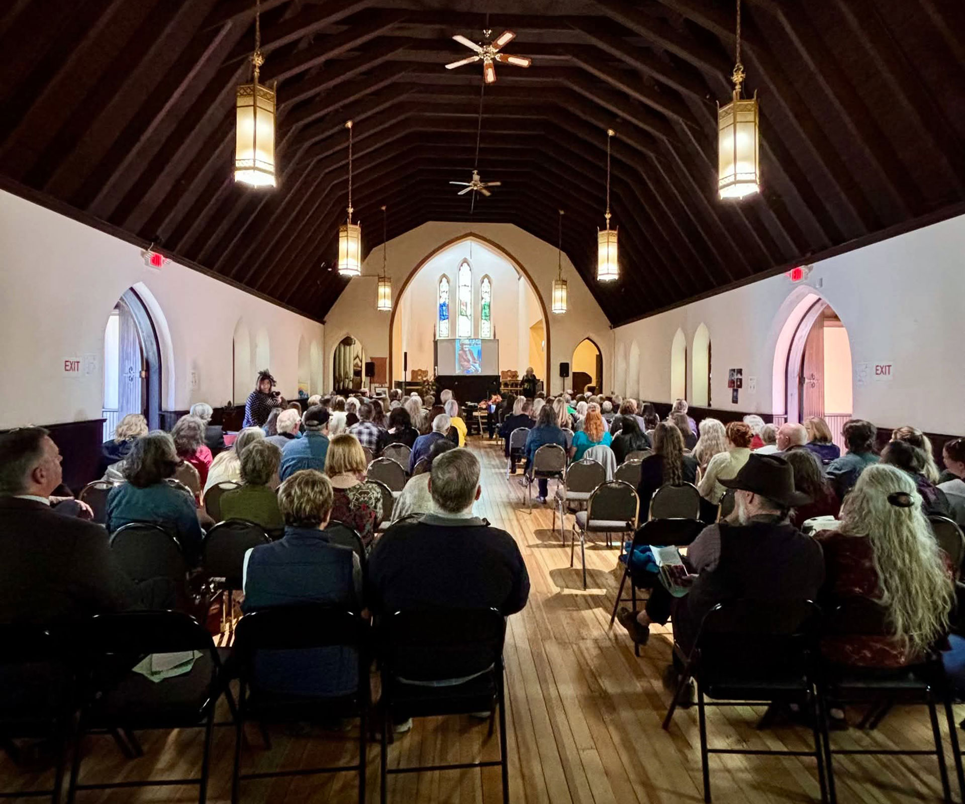 A packed community gathering inside the hall — rows of attendees facing the stained glass window at the far end.
